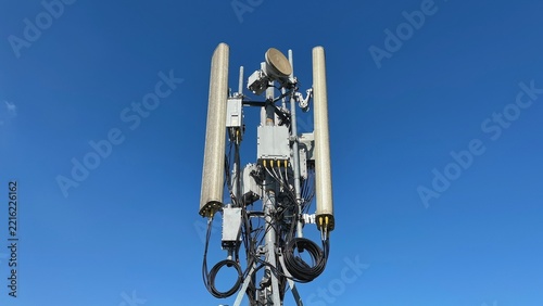 Clear blue sky with tall cellular tower structure. Multiple antennas and cables visible against the sky. Green foliage frames the bottom right corner. Ideal for telecom, infrastructure