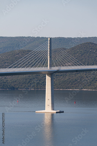 Symmetrical pylon of Peljesac Bridge reflected in calm bay, Croatia, clear day