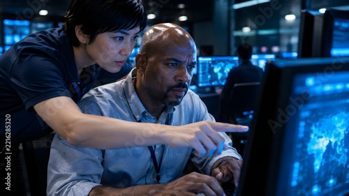 Two cybersecurity analysts collaborating in dark operations centre while pointing at world map threat monitor on computer screen