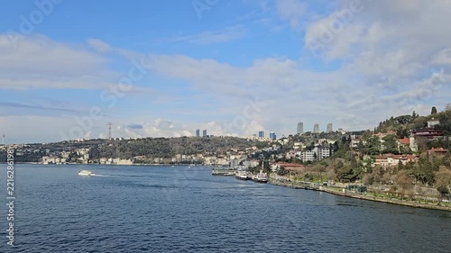 Blue sky above calm water with distant cityscape. White boat glides through the waterway. Hillsides covered with greenery and residential buildings in Istanbul, Turkey