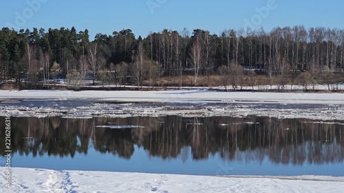 Frozen lake river reflects distant trees under clear sky. Snow blankets shorelines, ice patches float gently. Sky bright, blue, clouds absent above forested edge