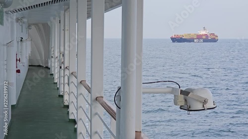 AMASRA - APR 16, 2026: Ship deck overlooks ocean. Large cargo vessel floats in distance. White railings line walkway. lamp mounted on cruise rail. Blue sea stretches wide. Sky is hazy and pale