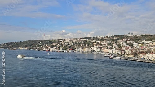 Boat glides across waters densely packed coastal towns. Sky holds clouds above hills and urban structures. Sailing vessel leaves gentle wake as it moves toward distant shoreline in Istanbul, Turkey