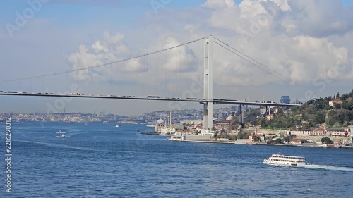 Grand bridge spans wide blue waters. Clouds drift above city skyline. Boats glide across calm sea surface. Vehicle traffic flows steadily. Distant hills rise behind bridge in Istanbul, Turkey