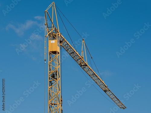 Yellow tower crane cabin and luffing jib boom against clean blue sky. Heavy construction machinery, housing development and real estate industry concept.