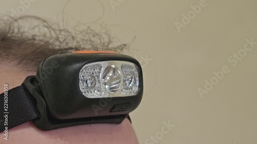 Red headlamp on person forehead. Black headband secures the lamp. Light shines brightly against plain wall. Person has dark, curly hair.