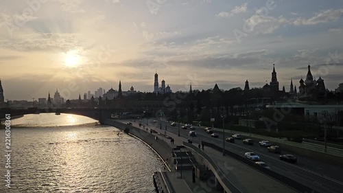 Sun sets over Moscow river and historic skyline. Silhouettes of spires stand against golden sky. River reflects sunlight as vehicles move along the road. People walk along the riverside path