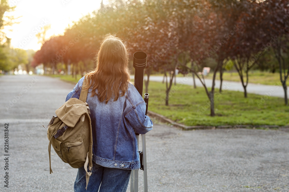 Fototapeta premium Astronomy and science education, young woman with telescope and backpack exploring nature and stargazing in a park during golden hour sunset