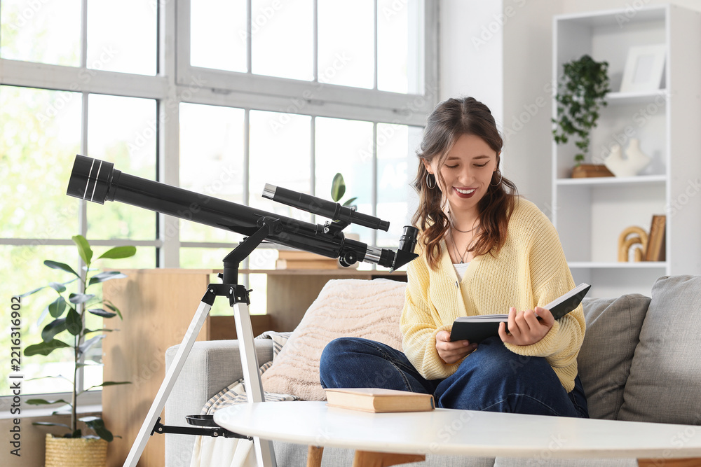Fototapeta premium Astronomy hobby and education, young woman reading a book about space science with a telescope at home in a bright living room
