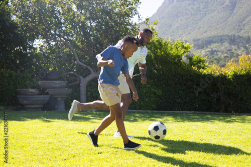 African American father and youth son playing soccer on garden lawn, wearing blue tee, kicking ball