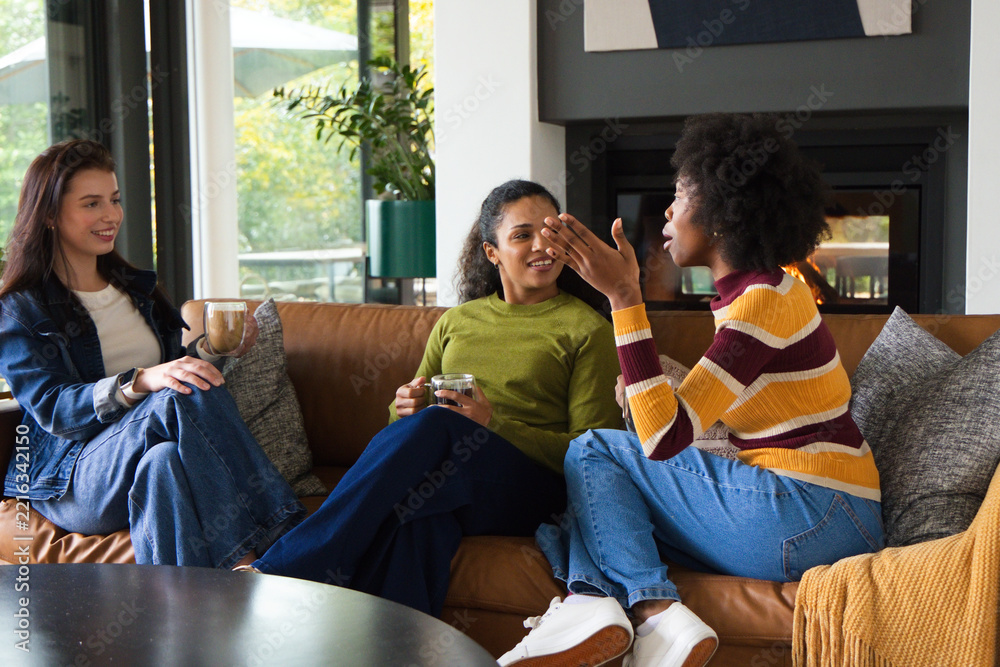 Fototapeta premium Diverse female friends sitting on brown leather sofa in living room, holding ceramic mug, glass cup