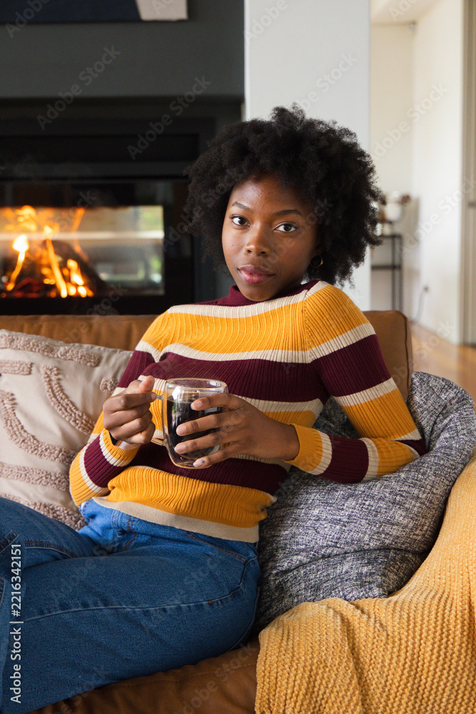 Fototapeta premium African American woman sitting at home on sofa wearing striped sweater holding glass mug near fire