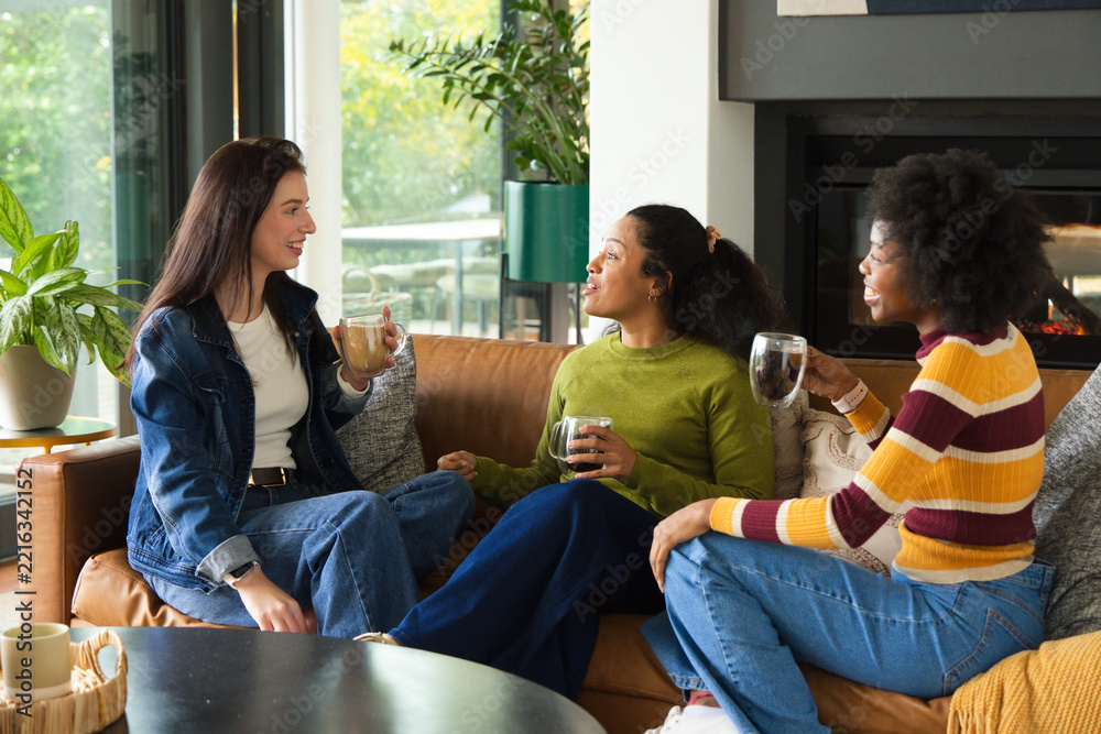 Fototapeta premium Diverse female friends sitting closely on leather sofa in modern living room holding mugs