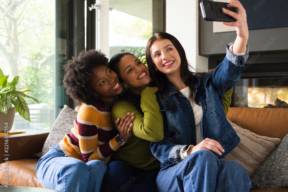 Fototapeta premium Diverse female friends posing for selfie on leather sofa in modern living room, holding smartphone