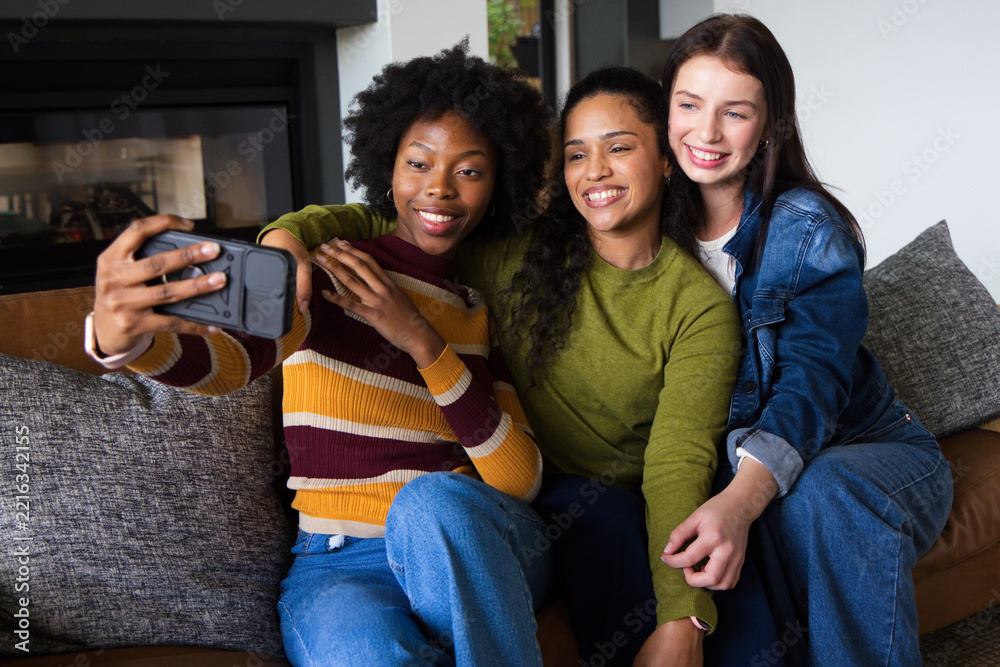 Fototapeta premium Diverse female friends sitting on sofa in living room taking selfie with smartphone case and jeans