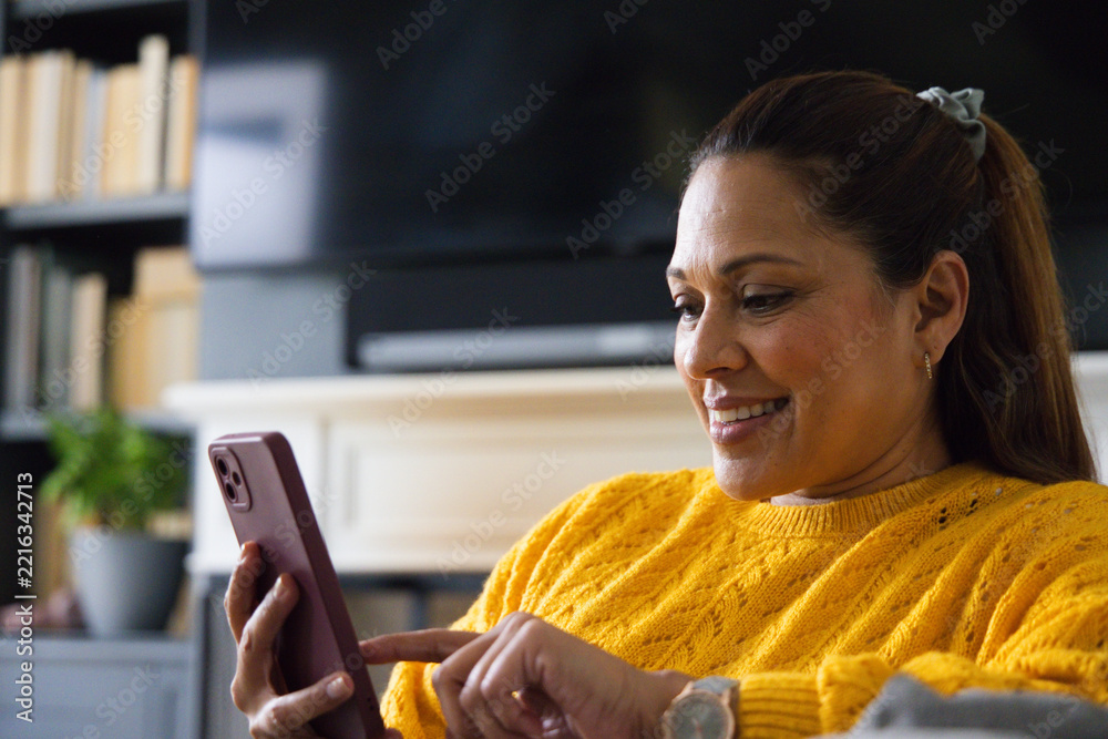 Fototapeta premium Middle-aged African American woman wearing mustard knit on sofa in living room tapping mauve case