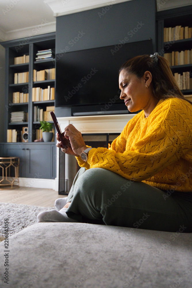 Fototapeta premium Mature woman sitting on low couch in living room wearing mustard-yellow sweater holding smartphone