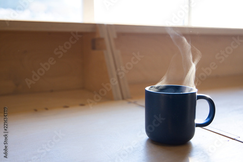 Dark blue ceramic mug is steaming on light wood countertop by window, soft sunlight, copy space