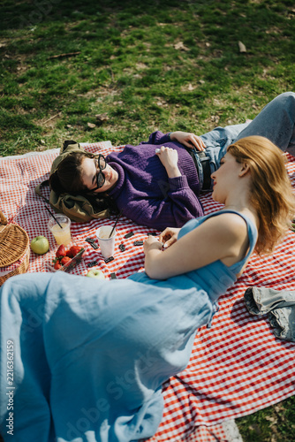Two young women lie on a red checkered picnic blanket enjoying a relaxed conversation. A casual outdoor picnic scene with fruit, drinks and leisure in the grass.