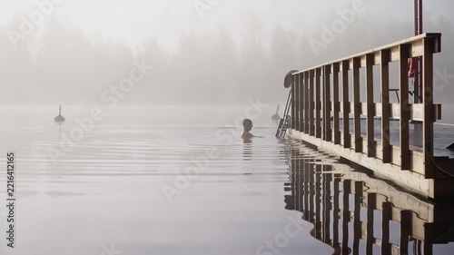 Middle aged man entering cold lake water from wooden dock early autumn morning foggy landscape Finland