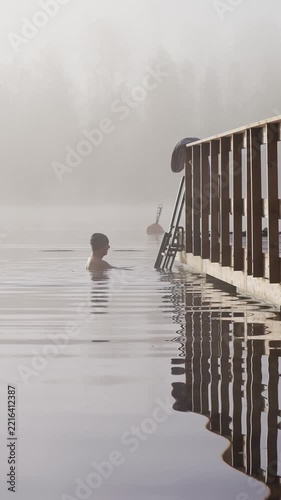 Middle aged man entering cold lake water from wooden dock early autumn morning foggy landscape Finland