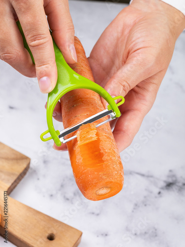 Woman peeling a carrot with a green-handled vegetable peeler on a marble-look countertop, close-up