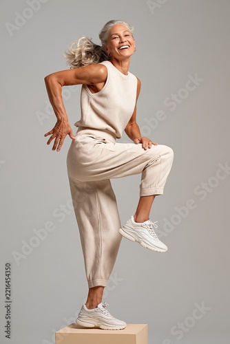 Fit older woman performing high knee exercise in studio with joyful expression and knit fresh outfit