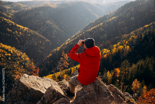 Man sitting on a rocky ledge and looking through binoculars at an autumn mountain landscape.