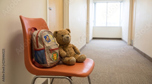 Small Child Rucksack And Worn Teddy Bear Waiting Together On Orange Plastic Chair In Corridor Showing Foster Care