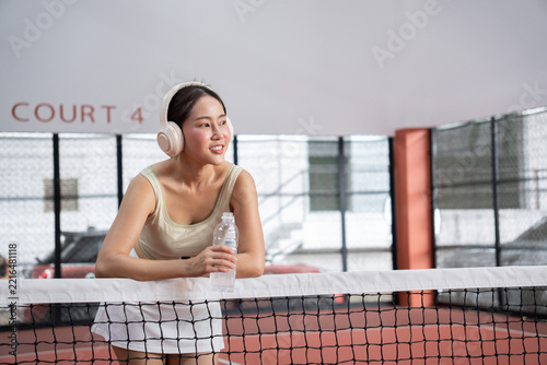 Obraz na plátně Asian woman wearing headphones holding water bottle and leaning on net taking a
