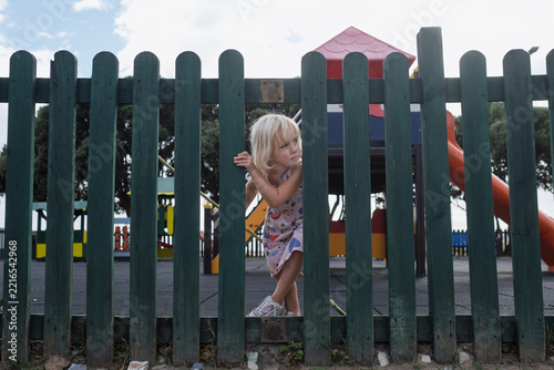 Little Girl Escaping and Squeezing Through Wooden Fence at Outdoor Playground