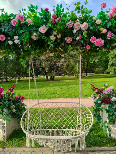 Beautiful park photo zone featuring woven macrame hammock swing, adorned with arch of lush greenery and pink garden roses. Sunny summer day.  Copy space.