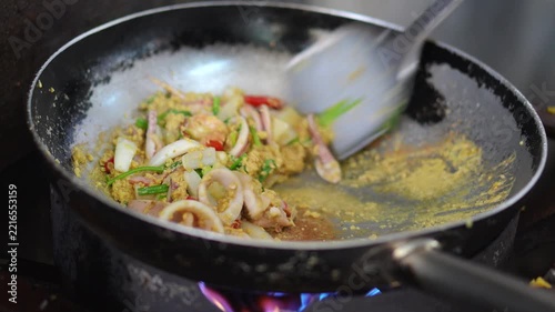 Chef using spatula to stir fry fresh squid rings with chili, garlic, and basil in a hot skillet over stove flame, showing cooking process for spicy Thai stir-fried squid concept.