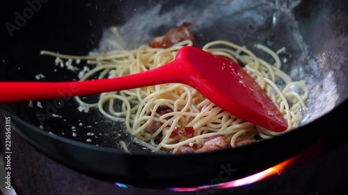 4K Chef using a red silicone spatula to stir fry spaghetti pasta with bacon in a hot wok over stove flame, cooking process for Italian spaghetti carbonara dish.