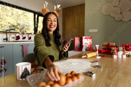 Smiling woman in reindeer antlers holding smartphone while selecting eggs for baking