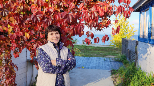 Mature woman standing under vibrant red autumn foliage near waterfront, wearing casual vest, smiling with crossed arms beside wooden house with sea view background, fall season lifestyle portrait