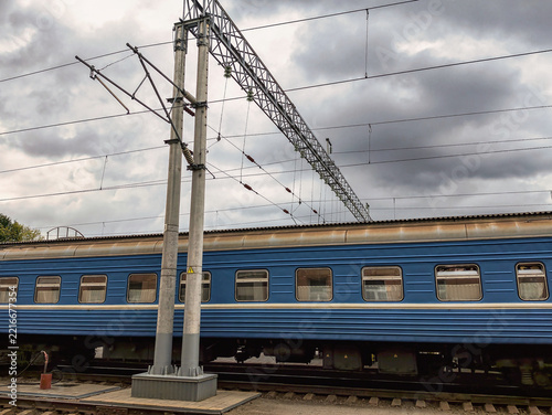 A blue train is parked at a train platform as dark clouds fill the sky. Overhead power lines are visible along the tracks at the station