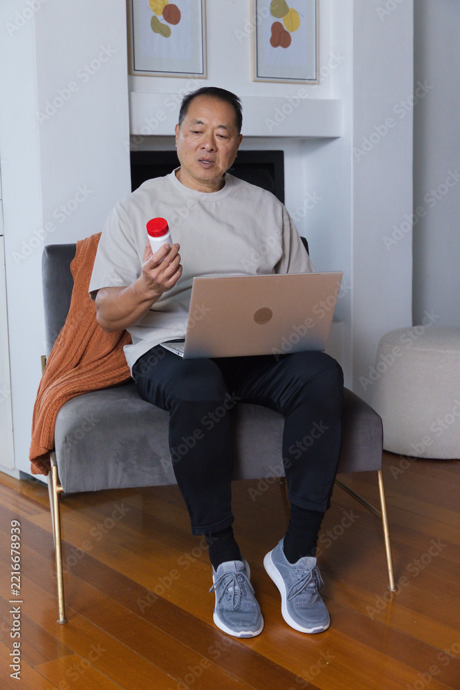 Naklejka premium Senior Asian man sitting in living room, reading white bottle label while using laptop