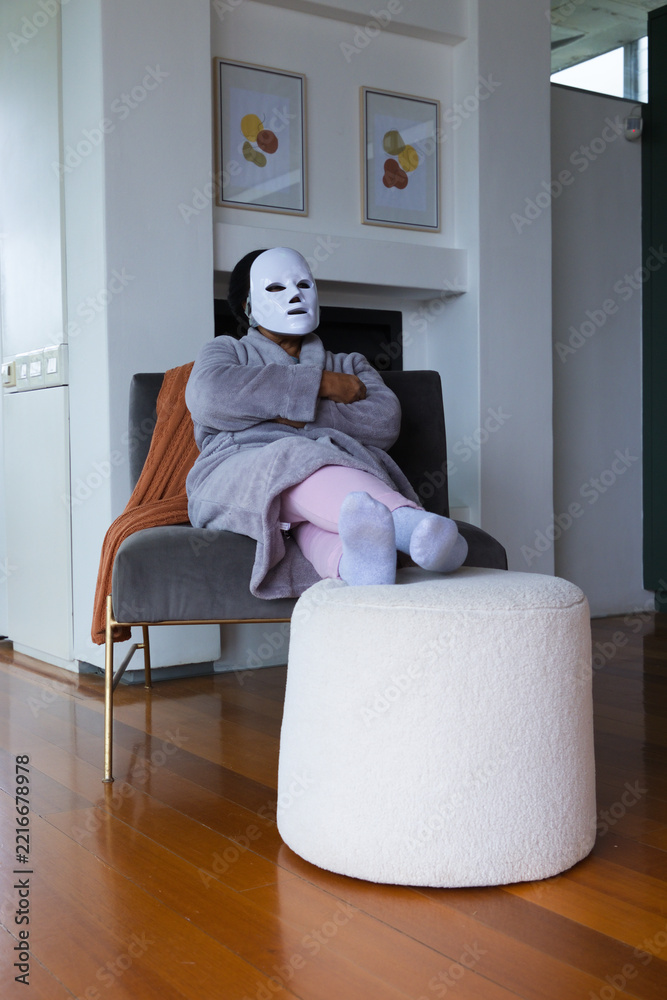 Fototapeta premium Senior African American woman sitting on lounge chair in living room wearing robe and white mask