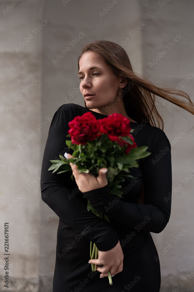 Fototapeta premium Vibrant floral portrait features woman in elegant dark attire holding bunch of red peonies against neutral wall outdoor setting