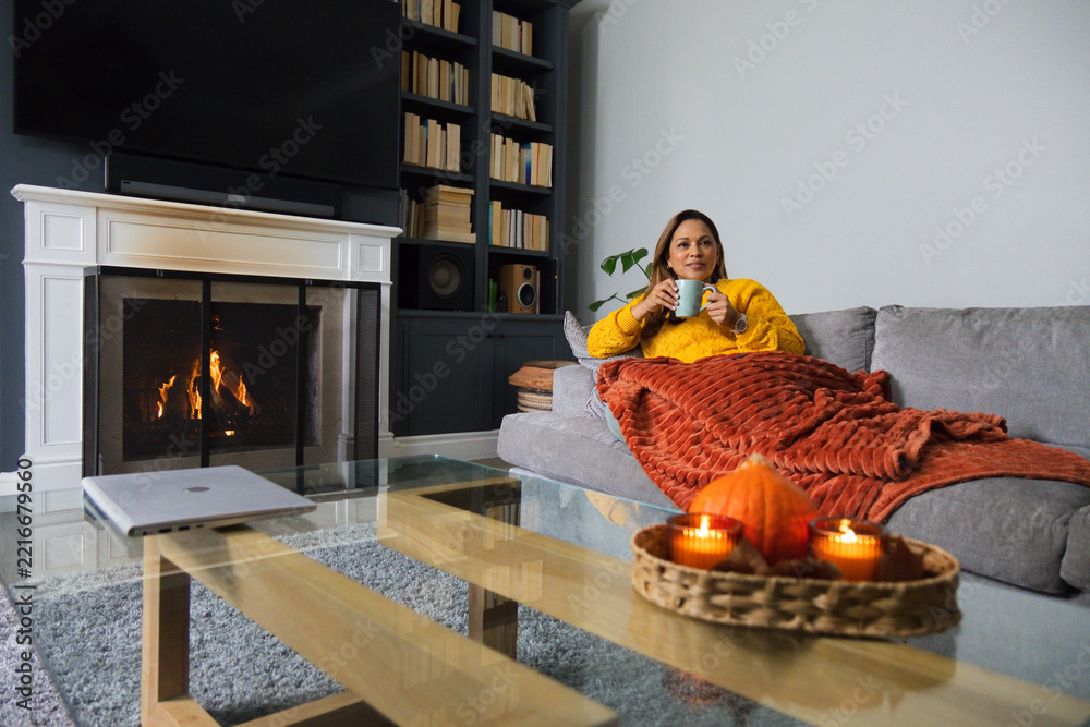 Fototapeta premium Mature African American woman lounging sofa in mustard-top holding blue mug by fireplace