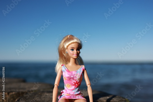 Fashion doll sitting on old wooden breakwater by the blue sea. Blonde toy doll in pink dress at summer beach. Childhood play and vacation concept.