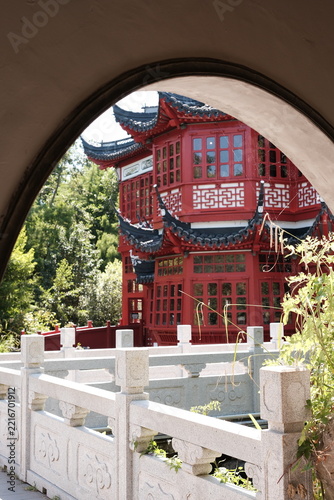 Traditional red Chinese pavilion with curved tiled roofs framed by a round moon gate arch, stone balustrade and green garden
Red wooden Chinese pavilion with dark grey tiled upturned eaves and carved 