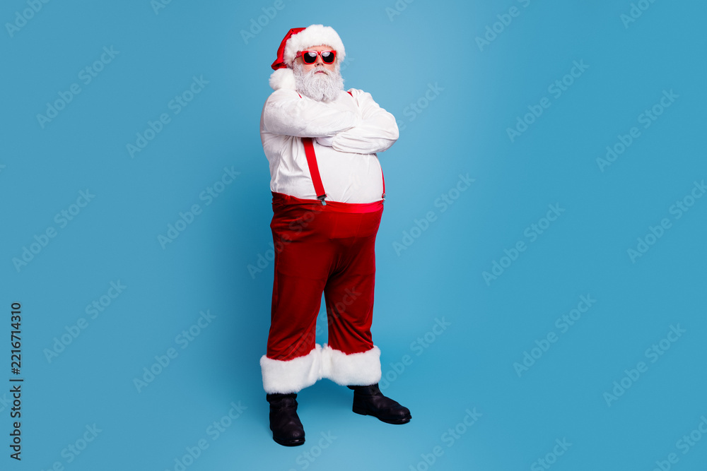 Fototapeta premium Cool Santa Claus standing with arms crossed in red suit and sunglasses on blue background, confident festive Christmas portrait