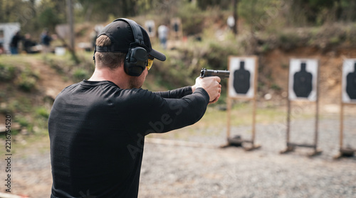 Rear View of Man in Black Shirt and Ear Protection Aiming a Handgun at Targets at an Outdoor Shooting Range. Concept of Firearm Training, Marksmanship, Safety Practice, and Sport Shooting.