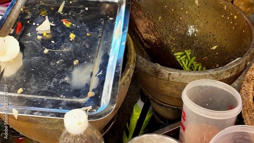 Close up shot of street food vendor hands pounding and mixing fresh cherry tomatoes with dark spicy sauce inside a clay mortar using a wooden pestle for Thai papaya salad.
