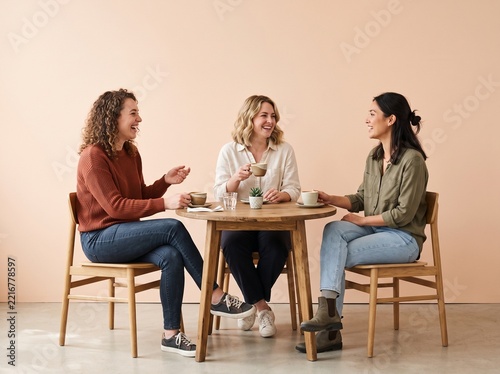 Three diverse female friends laughing and chatting while sitting at a round wooden table with coffee cups in a bright, modern studio setting with a soft peach background