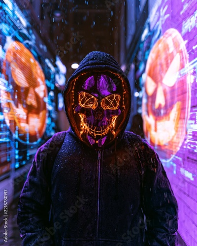 A figure in a dark hoodie and glowing mask stands in a Halloween alley