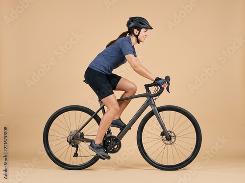 Side view of a young woman riding a modern road bike, wearing a cycling helmet and athletic clothing, studio shot against a solid beige background