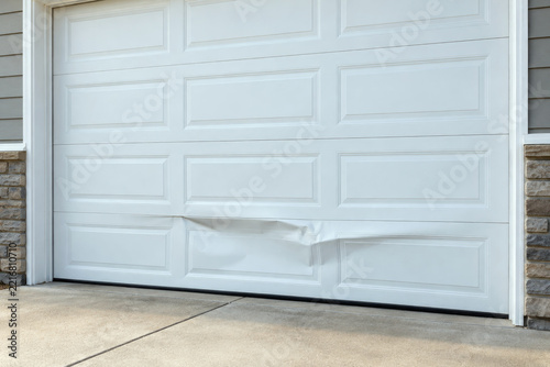 Dented and creased bottom panel on white residential door seen from driveway - garage door repair and panel replacement